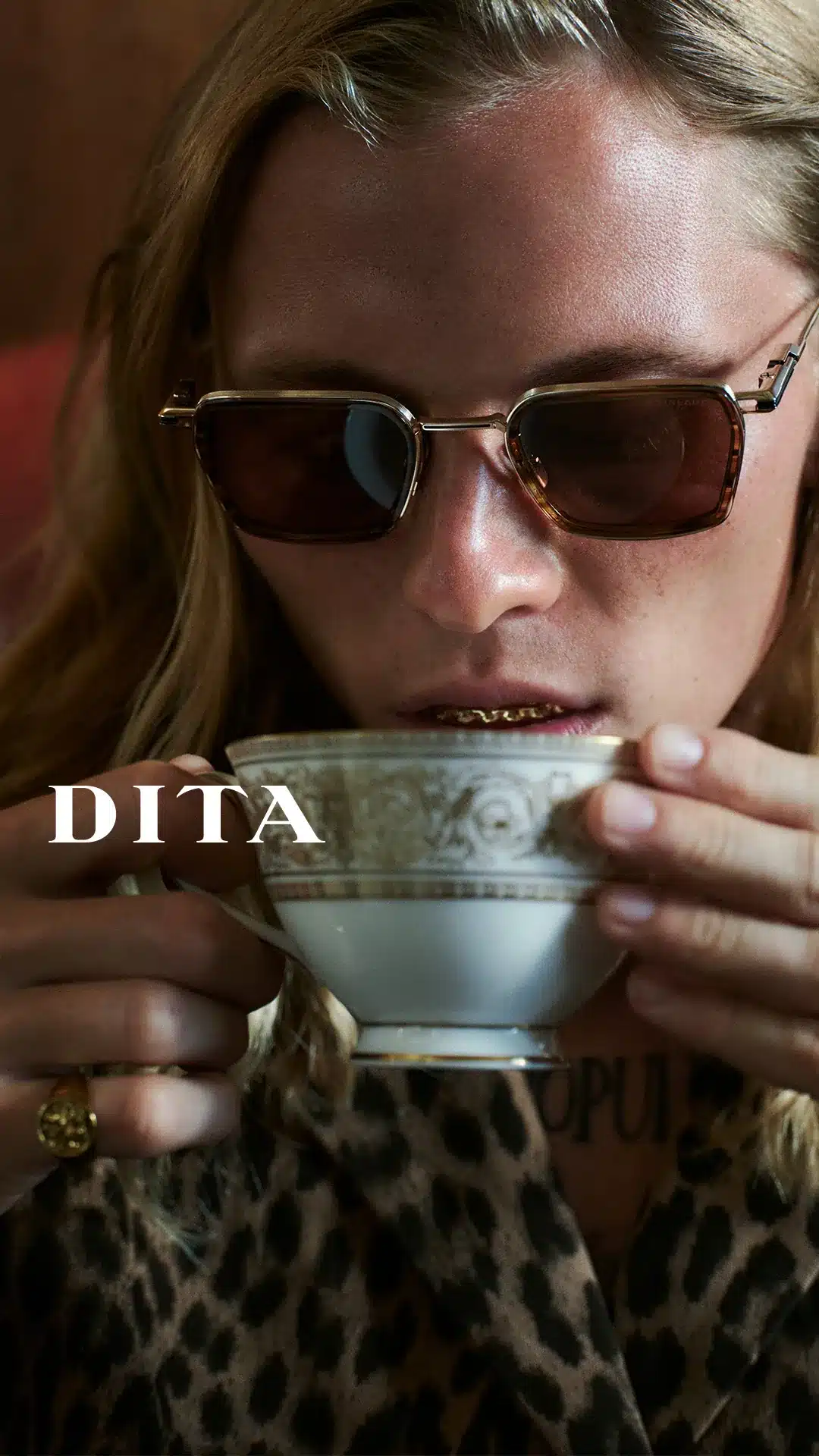 Model wearing DITA gold-rimmed sunglasses with tinted lenses, holding a porcelain teacup