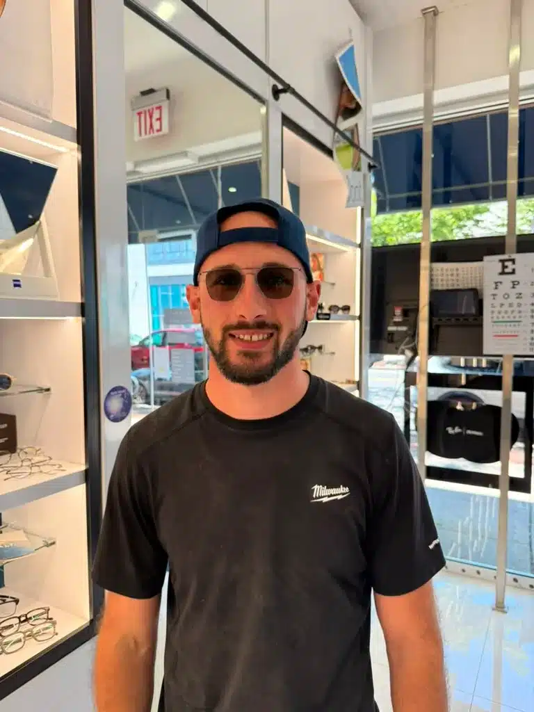 Man wearing tinted glasses and a backward black cap beside eyeglass display cases in an optical shop